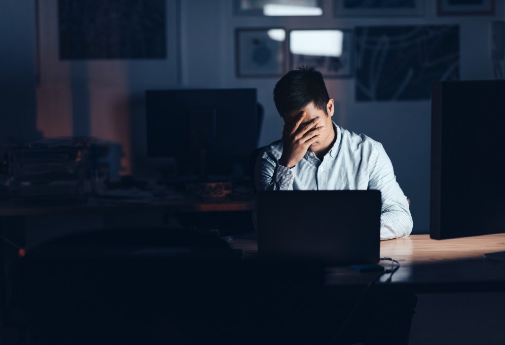 Young Asian businessman looking exhausted with his head in his hands while working alone at his desk in an office late at night